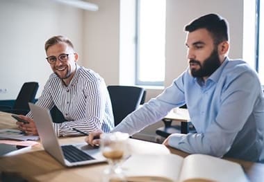 Two men sitting at a table with laptops discussing their pay.